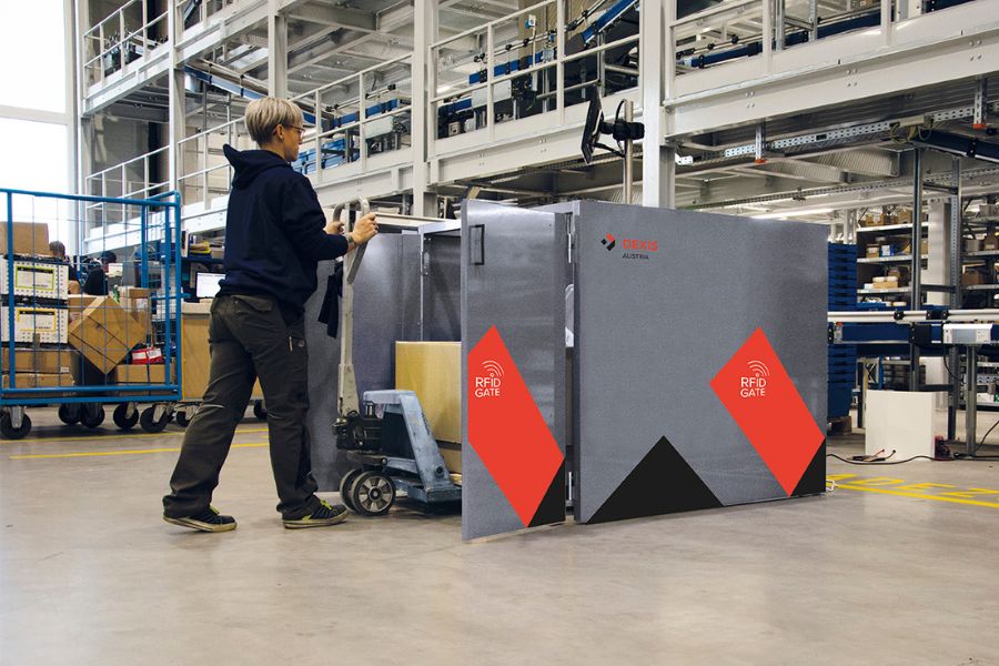 Eine dunkel gekleidete Mitarbeiterin schiebt die Palette aus dem RFID Gate in einem großen Industrielager mit Regalen, Förderbändern und verschiedenen Geräten im Hintergrund. Das RFID-Gate ist grau mit roten und schwarzen Akzenten.